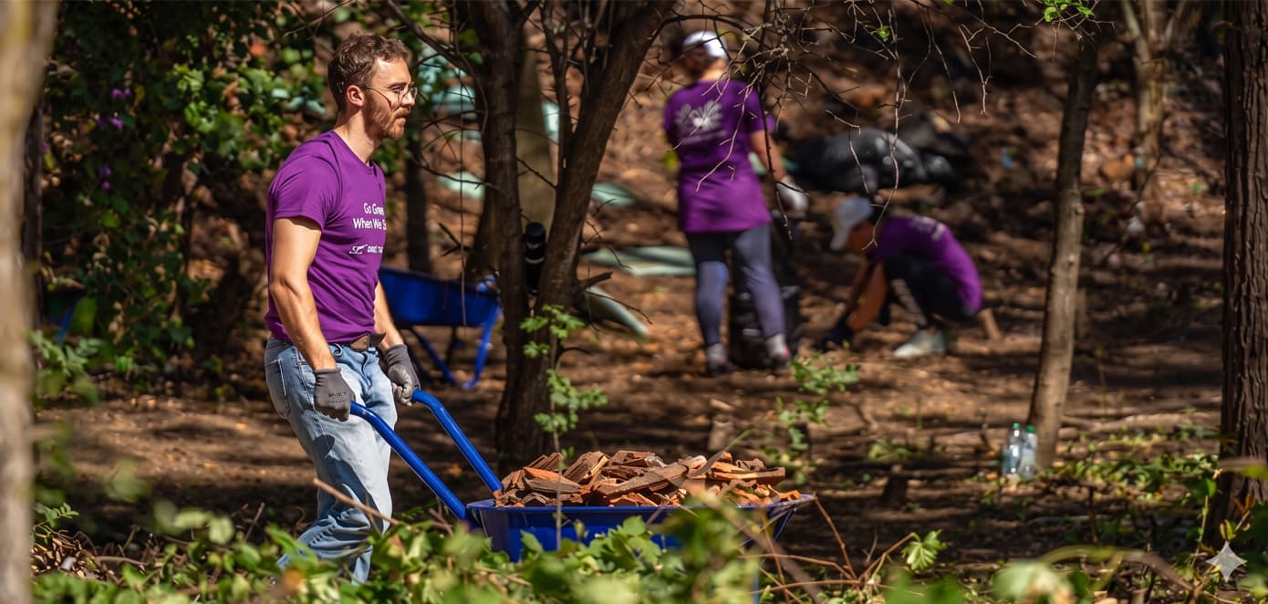 Several people wearing purple TELUS t-shirts work outside gardening. The person in the foreground is pushing a wheel barrow, while two people in the background are focusing on planting vegetation.