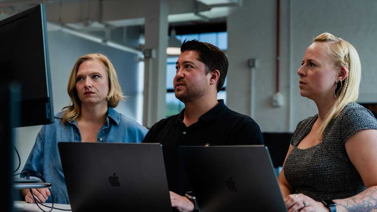 Three people sitting in front of laptops look up at something.